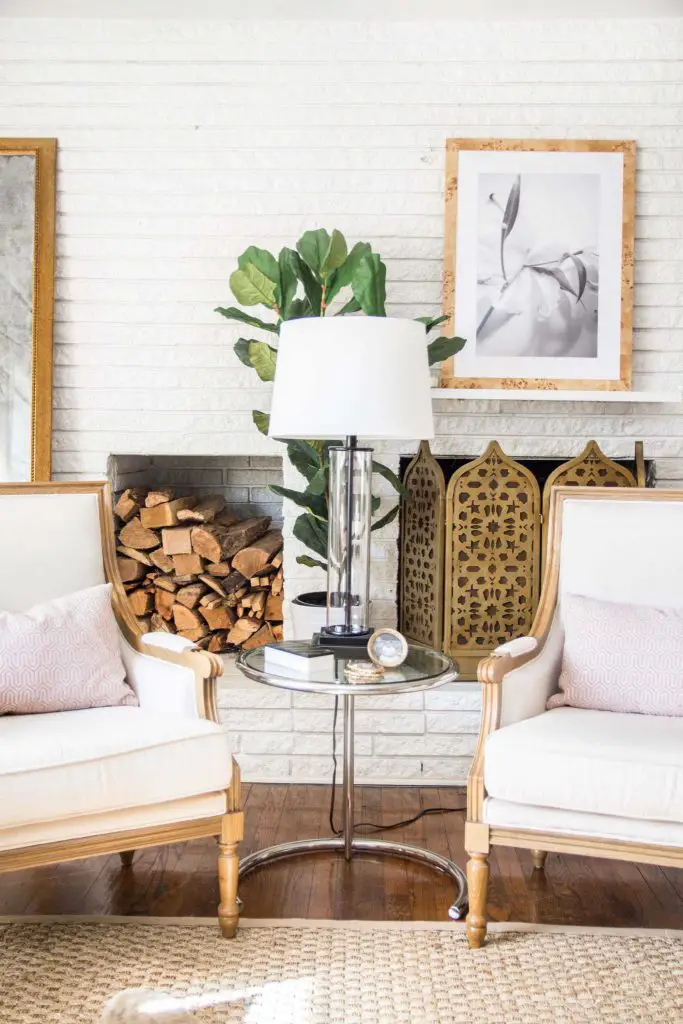 White brick fireplace with gold fireplace screen, stacked logs, linen armchairs, and pink pillows on Thou Swell #livingroom #livingroomdesign #homedecor #homedesign #interiordesign #pinkdecor #blush